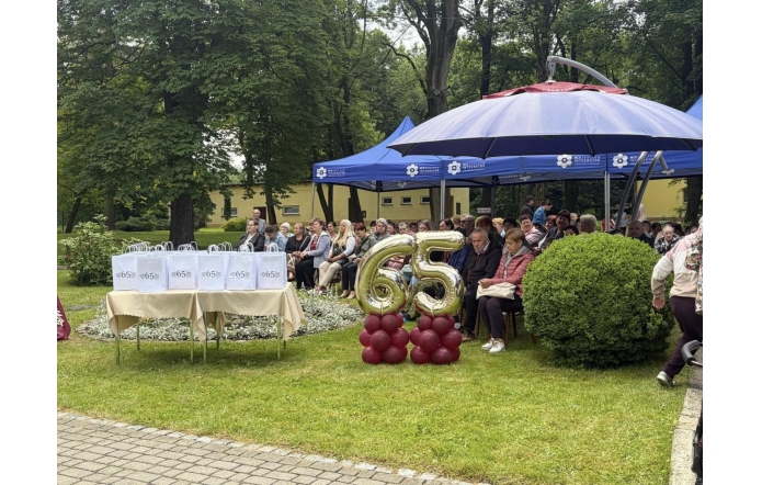 Publiczność siedzi w ogrodzie pod namiotami podczas uroczystości. Na pierwszym planie stoją złote balony z liczbą 65 oraz stolik z białymi torbami upominkowymi.