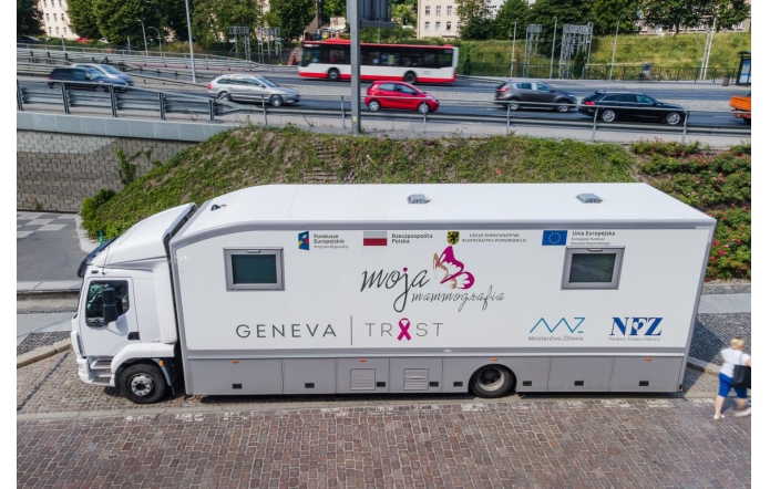 Biały mammobus stoi przy ulicy. Na jego boku widoczne są napisy i logotypy związane z programem „Moja Mammografia”, Funduszami Europejskimi, Unią Europejską, Ministerstwem Zdrowia i NFZ. W tle znajduje się ruchliwa jezdnia z samochodami i autobusem.