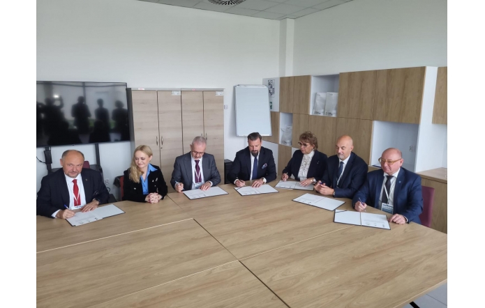 Osoby siedzą przy stole konferencyjnym i podpisują dokumenty. Na blacie widoczne są teczki oraz długopisy, a w tle znajdują się szafy, tablica i ekran.