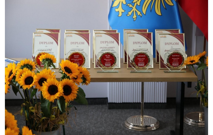Dyplomy i szklane statuetki ustawione na stole, przygotowane do wręczenia podczas uroczystości. Obok stoją bukiety słoneczników, a w tle widać flagi.