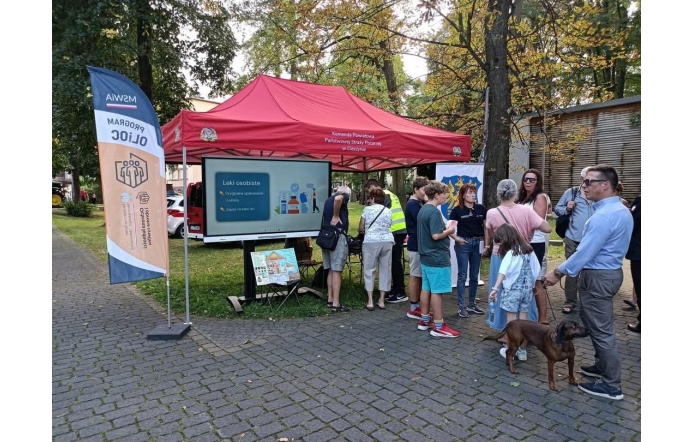 Grupa osób stoi przy czerwonym namiocie Komendy Powiatowej Państwowej Straży Pożarnej w Cieszynie . Pod namiotem znajduje się ekran z prezentacją dotyczącą leków osobistych oraz stół z materiałami informacyjnymi. Obok ustawiono flagę programu OLiOC, a na pierwszym planie stoją uczestnicy.