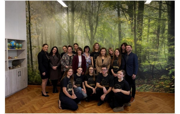 Grupa osób stoi i klęczy do wspólnego zdjęcia w pomieszczeniu z leśną grafiką na ścianie, obok widać regał z globusami.