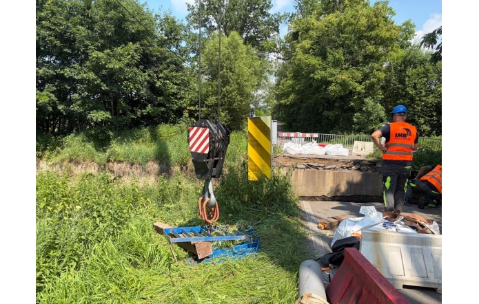 Prace przy uszkodzonym moście. Widoczny jest hak dźwigu, bariery zabezpieczające, materiały budowlane oraz pracownik w odzieży odblaskowej. Wokół znajduje się gęsta zieleń.