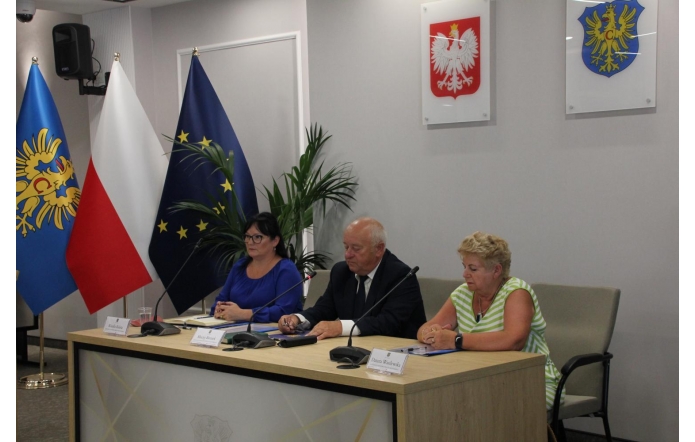 Trzy osoby siedzą przy stole prezydialnym w sali konferencyjnej. Kobieta w niebieskiej sukience, mężczyzna w garniturze i starsza kobieta w pasiastym topie. Za nimi flagi Polski, UE, województwa oraz herby na ścianie.