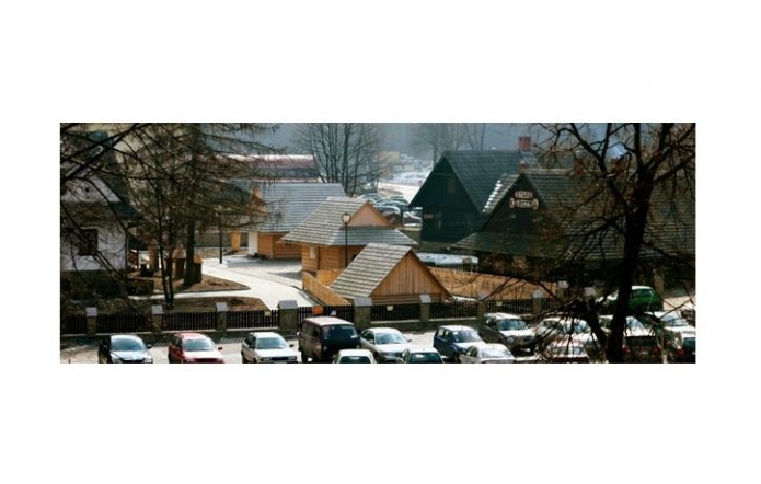 Zimowy widok drewnianych budynków Muzeum Beskidzkiego w Wiśle. Na pierwszym planie stoją zaparkowane samochody, a teren i dachy są częściowo pokryte śniegiem.