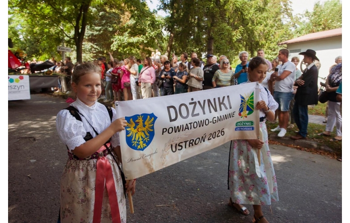 Dwie dziewczynki w strojach regionalnych niosą baner z napisem „Dożynki Powiatowo-Gminne Ustroń 2026”. W tle stoją licznie zgromadzeni uczestnicy i widzowie korowodu.