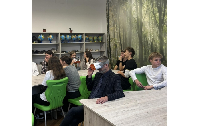 Uczniowie siedzą przy stołach w pracowni z zielonymi krzesłami. Michał Rajwa Naczelnik Wydziału Edukacji korzysta z gogli VR, a w tle widoczne są globusy, minerały oraz fototapeta z motywem lasu.