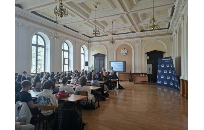 Uczniowie siedzą przy ławkach w dużej sali. Z przodu znajdują się osoba prowadząca, ekran z prezentacją oraz baner „Szkoła dla ludzi z pasją”.