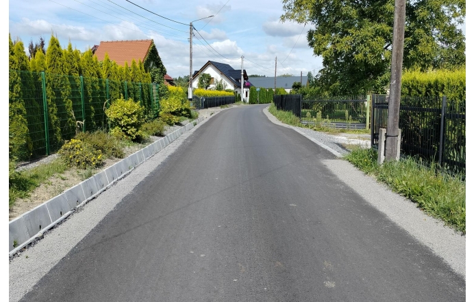 Wyasfaltowana droga biegnie między ogrodzeniami posesji z zielenią i krzewami. W tle widać domy jednorodzinne, a nad nimi niebo z białymi chmurami.