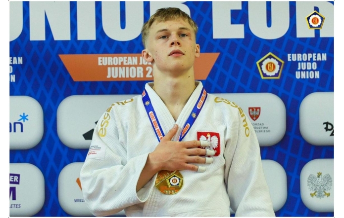 Młody judoka w białym stroju stoi na podium z medalem na szyi, trzymając rękę na piersi. W tle widoczna jest ścianka.