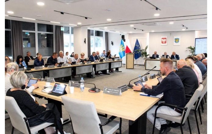 Widok na salę sesyjną podczas obrad. Uczestnicy siedzą przy ustawionych w podkowę stołach, przed nimi znajdują się mikrofony, tablety i tabliczki z nazwiskami. W centralnej części sali widoczne są mównica oraz flagi, a na ścianie godło Polski i herb Powiatu Cieszyńskiego.