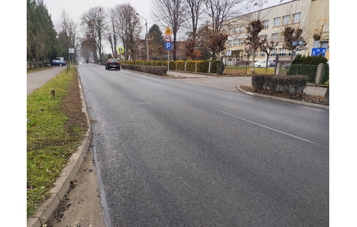 Droga asfaltowa z chodnikiem i znakami drogowymi. W tle widać samochody, drzewa oraz budynek za ogrodzeniem.
