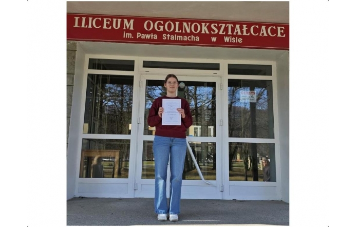 Dziewczyna stoi przed wejściem do liceum i trzyma w rękach dyplom, nad drzwiami widnieje napis z nazwą szkoły w Wiśle.