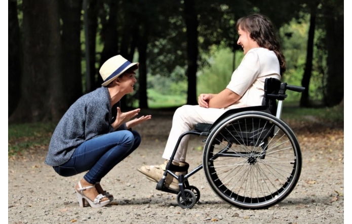 Dwie kobiety rozmawiają w parku. Jedna siedzi w wózku inwalidzkim, ubrana na jasno, uśmiecha się. Druga kuca naprzeciw niej, gestykuluje radośnie, nosi kapelusz, jeansy i buty na obcasie. Obie wyglądają na szczęśliwe.