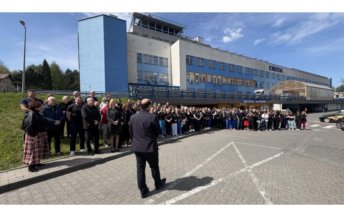 Duża grupa ludzi stoi przed budynkiem i słucha mężczyzny przemawiającego do nich na zewnątrz, w słoneczny dzień przy wejściu głównym.