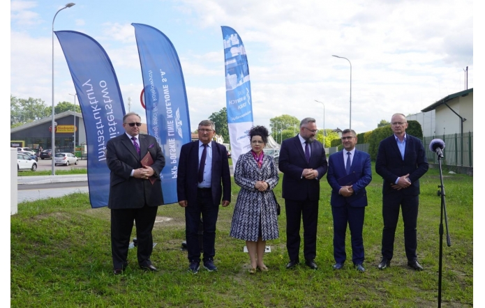 Sześć osób stoi na trawie przy drodze. Obok nich ustawione są niebieskie flagi promocyjne oraz mikrofon na statywie. W tle widać ulicę, samochody i budynki.