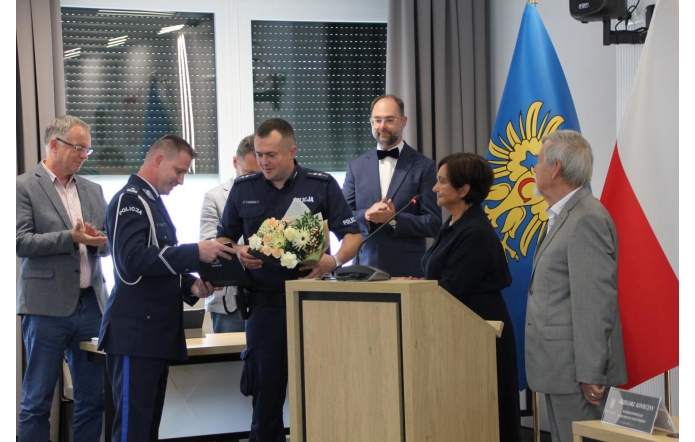 Podczas uroczystości w sali sesyjnej dwóch policjantów odbiera gratulacje i kwiaty z rąk przedstawicieli władz. Obok mównicy stoi Janina Żagan, a w tle widoczne są flagi: województwa śląskiego i Polski.