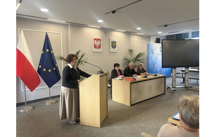 Sala sesyjna, Przy mównicy stoi Janina Żagan Starosta Cieszyński, ubrana elegancko, przemawia do kilku osób siedzących przy stole. W tle flagi i ekran projekcyjny. Na stole mikrofony i dokumenty.