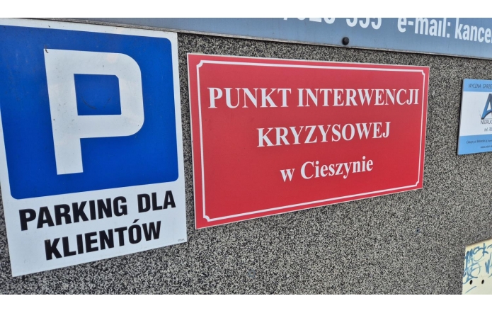 Zdjęcie przedstawia dwa znaki na ścianie budynku. Jeden to niebieski znak parkingowy z literą „P” i napisem „PARKING DLA KLIENTÓW”. Drugi to czerwona tablica z białym napisem „PUNKT INTERWENCJI KRYZYSOWEJ w Cieszynie”, oznaczająca miejsce pomocy.