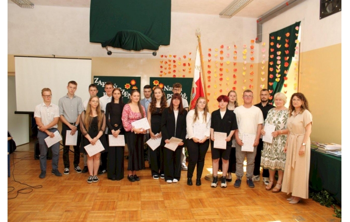 Grupa uczniów i nauczycieli pozuje wspólnie do zdjęcia podczas uroczystości zakończenia roku szkolnego. W tle kolorowa dekoracja z motylkami, sztandar oraz plansze z napisem „Zakończenie roku”.
