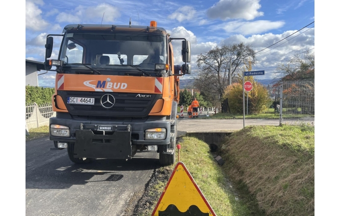Pomarańczowy pojazd budowlany stoi na remontowanej drodze. Obok widoczne są znaki drogowe, fragment pobocza oraz pracownicy w odblaskowej odzieży w głębi ulicy.