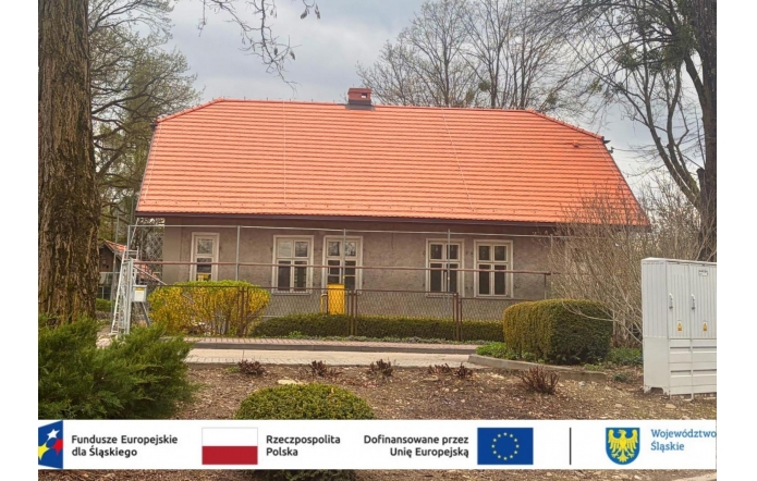 Budynek z nowym czerwonym dachem i odnowioną elewacją, otoczony ogrodzeniem i niską zielenią; przed nim chodnik i krzewy, a u dołu widoczny pasek z logotypami i informacją o dofinansowaniu z funduszy europejskich.