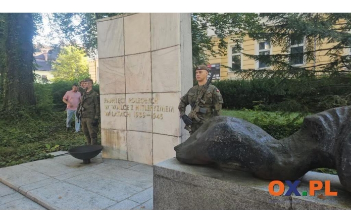 Pomnik z jasną kamienną płytą z napisem oraz ciemną rzeźbą umieszczoną na cokole. Po obu stronach monumentu stoją żołnierze w mundurach, a w tle widoczne są drzewa, krzewy i budynek.