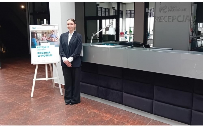  Ewa Raszka w eleganckim stroju hotelowym stoi przy recepcji Term Warmińskich. Obok znajduje się plakat promujący program "Rodzina w hotelu" organizowany przez SGH. Scena oddaje atmosferę profesjonalizmu i gościnności