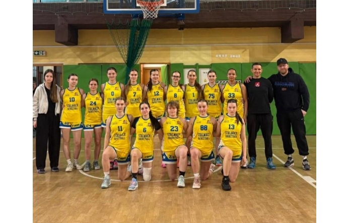 Grupa zawodniczek w żółtych strojach koszykarskich oraz trener na sali gimnastycznej pod koszem do gry.