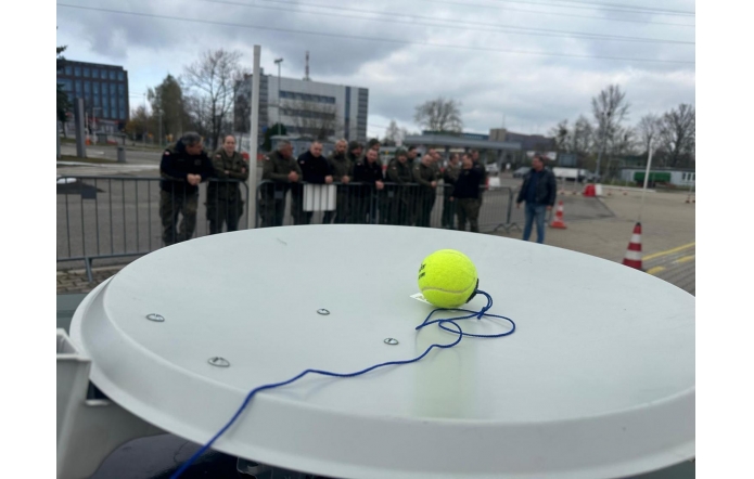 Żółta piłka tenisowa z przywiązanym niebieskim sznurkiem leży na białej antenie satelitarnej. W tle grupa umundurowanych osób stoi za barierką na placu, obok widoczne są pachołki drogowe i biurowce.