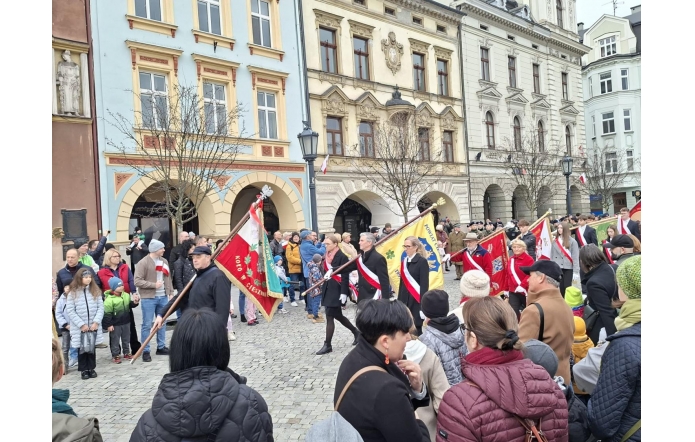 Uczestnicy przechodzą przez rynek z pocztami sztandarowymi. Wokół stoją zgromadzeni mieszkańcy, a w tle widoczne są zabytkowe kamienice.