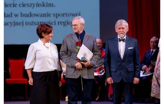Trzy osoby stoją na scenie podczas uroczystości. Mężczyzna pośrodku trzyma różę, dokument oraz małe pudełko, a w tle widoczne są czerwone kurtyny i ekran z tekstem.
