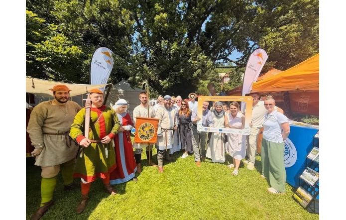 Grupa osób pozuje na trawniku podczas plenerowego wydarzenia. Część osób ma historyczne stroje i rekwizyty, a w tle widać namioty, flagi „Śląsk Cieszyński” oraz stoisko z materiałami promocyjnymi.