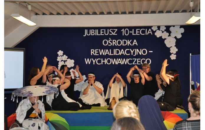 Grupa osób występuje na scenie podczas jubileuszu 10-lecia Ośrodka Rewalidacyjno-Wychowawczego. Część uczestników siedzi lub klęczy i wykonuje gesty rękami, a obok stoją osoby wspierające występ. W tle widoczny jest granatowy napis jubileuszowy oraz białe dekoracje kwiatowe.