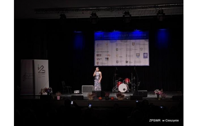 Młoda kobieta śpiewa na oświetlonej scenie podczas wydarzenia artystycznego; w tle widać baner sponsorów, zestaw perkusyjny oraz dekoracje ustawione.