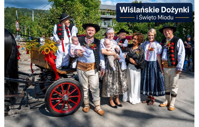 Grupa dorosłych i dzieci pozuje do wspólnego zdjęcia obok udekorowanego wozu zaprzęgowego. Część osób ma na sobie tradycyjne stroje regionalne, a niektórzy trzymają dzieci na rękach. W tle widoczne są drzewa, zabudowania oraz napis „Wiślańskie Dożynki i Święto Miodu”.