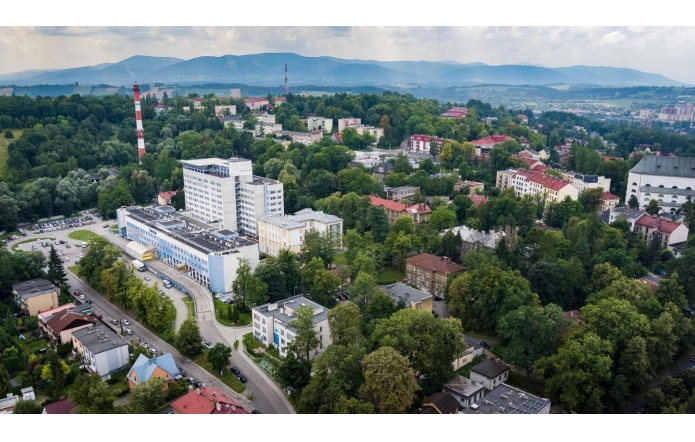 Widok z lotu ptaka na zabudowaną, zieloną okolicę Cieszyna z dużym kompleksem Szpitala Śląskiego w centrum kadru. W tle widać wzgórza, lasy, bloki mieszkalne i charakterystyczny komin w biało-czerwone pasy.