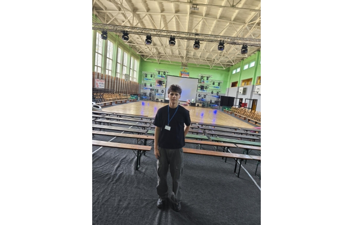 Mężczyzna stoi w centrum hali sportowej. W tle widoczne są ławki.