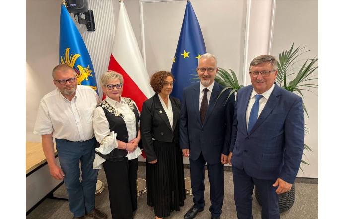 Zarząd Powiatu Cieszyńskiego pozuje do zdjęcia w sali przy flagach. W tle widać roślinę oraz fragment stołu.