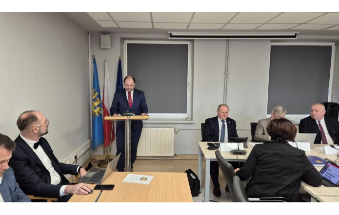 W Sali konferencyjnej Starostwa odbywa się XIX sesja Rady Powiatu Cieszyńskiego. Adam Śliż, dyrektor Szpitala Śląskiego w Cieszynie, ubrany w granatowy garnitur i czerwony krawat, stoi przy mównicy, przemawiając do obecnych. W tle widoczne są flagi narodowe oraz urzędowe. Przy stołach zasiadają radni w eleganckich strojach, skupieni na prezentowanych treściach. Atmosfera formalna i poważna.