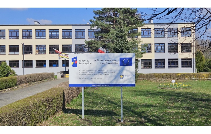Przed budynkiem szkoły stoi tablica informacyjna z logo Funduszy Europejskich i Unii Europejskiej, zawierająca informacje o dofinansowaniu projektu.