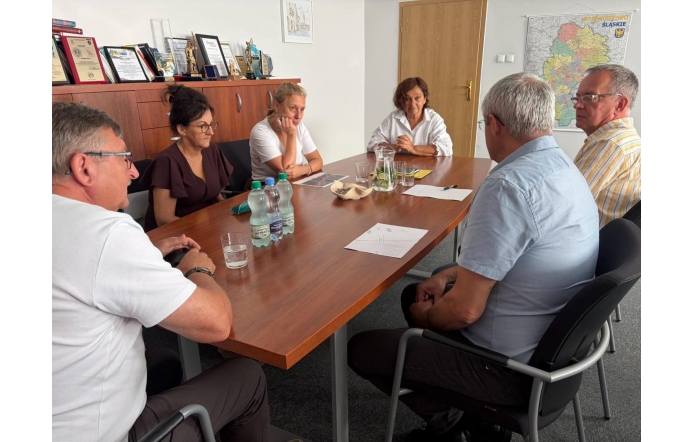 Grupa osób siedzi przy stole. Na stole widać dokumenty, szklanki, dzbanek i butelki z wodą, a w tle szafkę z dyplomami, statuetkami oraz mapę.
