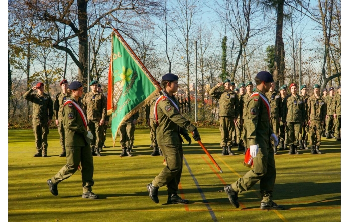 Grupa umundurowanych osób maszeruje na boisku podczas uroczystości. Na pierwszym planie widoczny jest poczet ze sztandarem, a w tle stoją kolejne osoby w szyku.