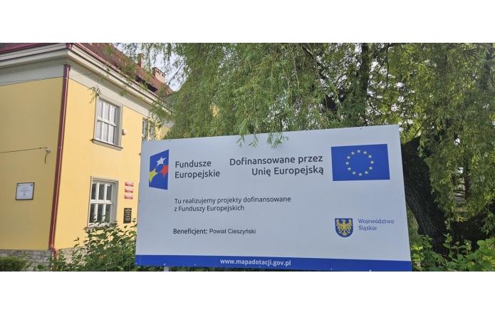 Tablica informacyjna o projekcie dofinansowanym przez Unię Europejską, ustawiona przed budynkiem i drzewami. Na tablicy widoczne są logotypy.