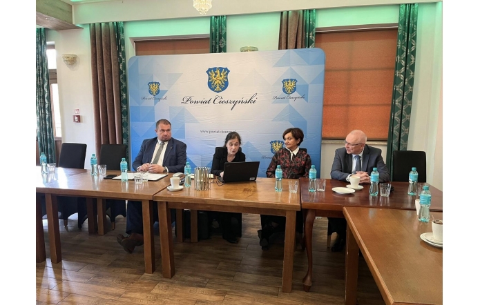 Sala konferencyjna z ustawionymi stołami. Przy stole siedzą cztery osoby, przed nimi znajdują się butelki z wodą, szklanki i laptop. W tle widoczna jest ścianka z herbem oraz napisem „Powiat Cieszyński”.