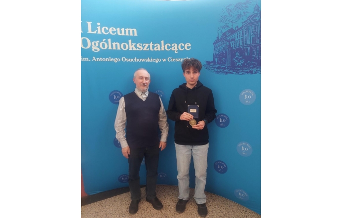 Dwóch mężczyzn na tle niebieskiej ścianki z napisem liceum; młodszy trzyma medal.