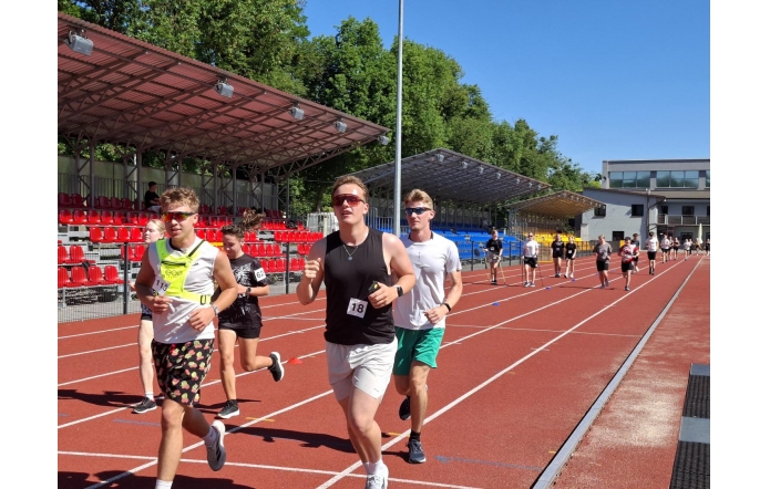 Grupa młodzieży biegnie po czerwonej bieżni lekkoatletycznej na stadionie. W tle widać trybuny z czerwonymi krzesełkami, drzewa oraz słoneczne niebo.