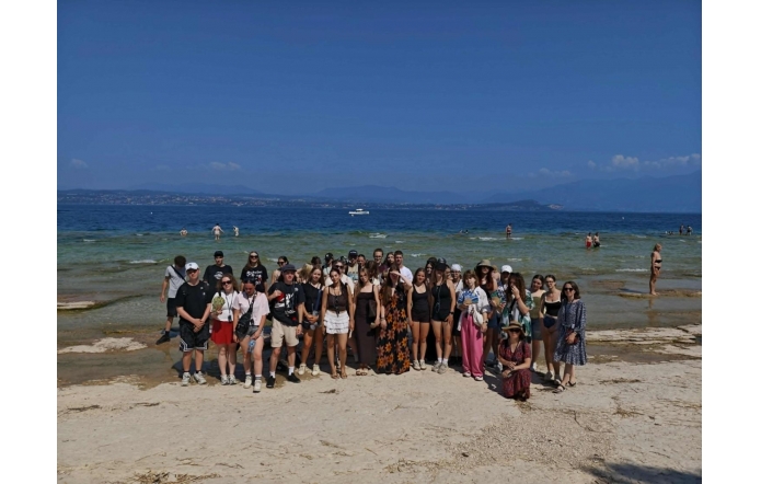 Grupa osób pozuje do zdjęcia na plaży. W tle widać wodę, kąpiących się ludzi, odległy brzeg oraz góry pod błękitnym niebem.