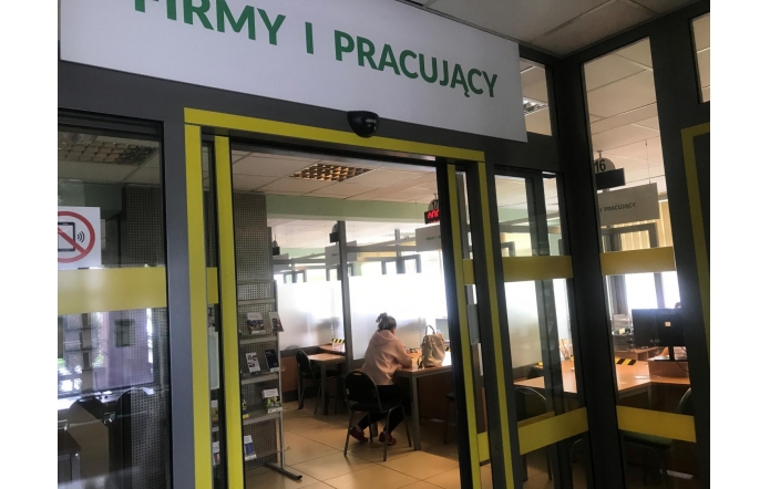 Pomieszczenie z oznaczeniem „FIRMY I PRACUJĄCY”, w którym osoby siedzą przy stanowiskach obsługi. Przestrzeń jest przeszklona, a w środku widoczne są biurka, krzesła i materiały informacyjne.