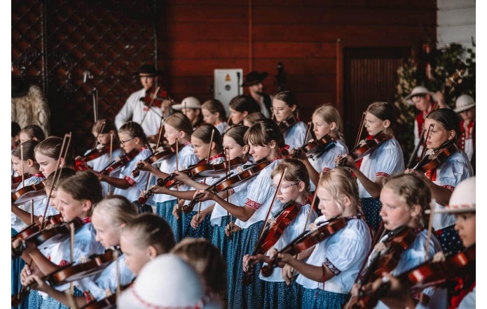 Dzieci i młodzież w strojach regionalnych grają na skrzypcach podczas występu. W tle widać muzyków prowadzących oraz drewnianą scenę.